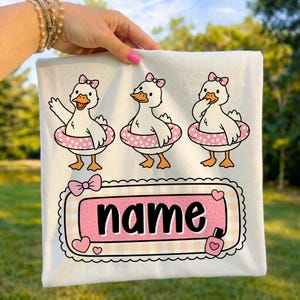 Puede incluir: Camiseta blanca con tres patos de dibujos animados con flotadores rosas de lunares y lazos. Debajo, un diseño de cuadros rosas y blancos con la palabra "name" en negro, enmarcada por corazones y un lazo.