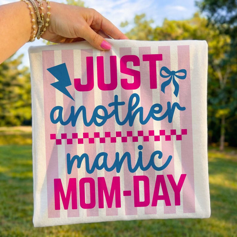 A close-up shot of a hand with pink manicured nails holding up a folded light blue t-shirt against a blurry green park background. The shirt features a vibrant graphic that reads Just Another Manic Mom-Day in a mix of bold pink and blue lettering. The design is set against a light pink and white striped box, accented with a blue lightning bolt, and a pink checkered line. This mockup highlights the crisp, colorful print quality and is perfect for showcasing funny mom-themed apparel for summer.