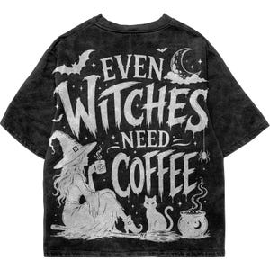 Può includere: Una t-shirt nera lavata all'acido con una stampa grafica bianca raffigurante una strega seduta su una scopa che tiene una tazza di caffè, un gatto, un calderone, una luna crescente e pipistrelli. Il testo recita "EVEN WITCHES NEED COFFEE".
