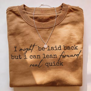 Brown folded t-shirt displayed with a necklace accent, featuring handwritten-style quote “I might be laid back but I can lean forward real quick,” centered chest print, warm tones, modern lifestyle flat lay presentation.