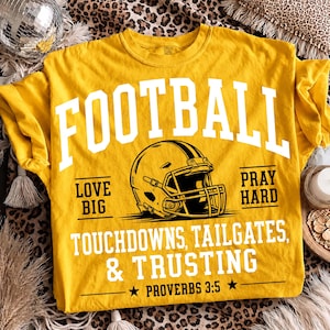 May include: A mustard yellow t-shirt featuring a white graphic of a football helmet. The text reads FOOTBALL, LOVE BIG, PRAY HARD, TOUCHDOWNS, TAILGATES, & TRUSTING, and PROVERBS 3:5. The design includes black and white lettering on the front.