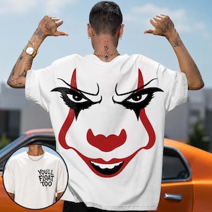 May include: A white short-sleeved t-shirt featuring a large graphic of a clown face with red and black details on the back. The front chest area displays the black text "YOU'LL FLOAT TOO" in a stylized font. The shirt has a crew neckline.