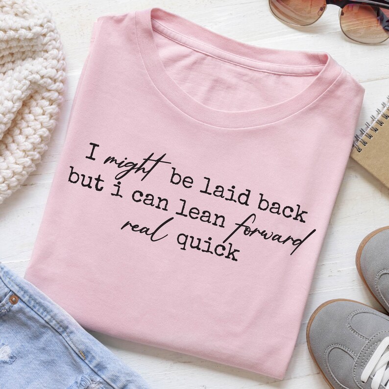 Pink folded t-shirt flat lay styled with denim and cozy accessories, showcasing minimalist handwritten quote “I might be laid back but I can lean forward real quick,” clean centered design, soft pastel aesthetic for casual wear.