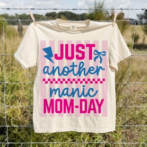 A light cream-colored t-shirt pinned with wooden clothespins to a wire fence in a sunny, rural outdoor setting. The graphic shows the Manic Mom-Day design in vivid pink and blue. The natural outdoor lighting showcases the soft texture of the Comfort Colors cotton fabric and the vibrancy of the colors. This rustic, nostalgic mockup is perfect for highlighting the shirt as a comfortable, durable, and breathable option for busy moms during summer outings or park days.