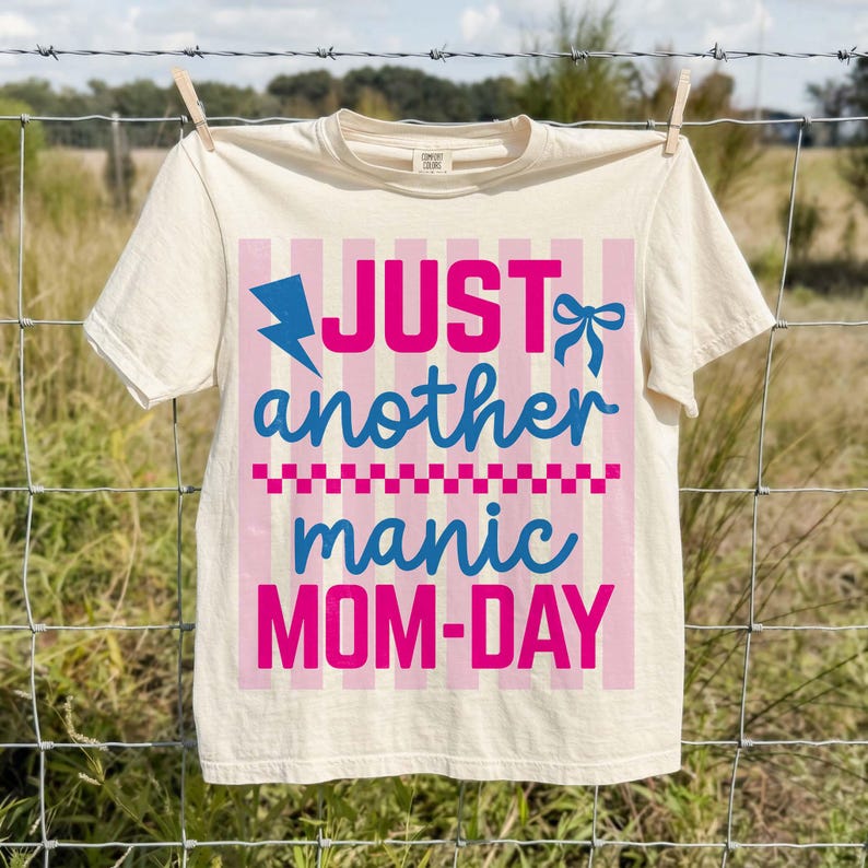 A light cream-colored t-shirt pinned with wooden clothespins to a wire fence in a sunny, rural outdoor setting. The graphic shows the Manic Mom-Day design in vivid pink and blue. The natural outdoor lighting showcases the soft texture of the Comfort Colors cotton fabric and the vibrancy of the colors. This rustic, nostalgic mockup is perfect for highlighting the shirt as a comfortable, durable, and breathable option for busy moms during summer outings or park days.