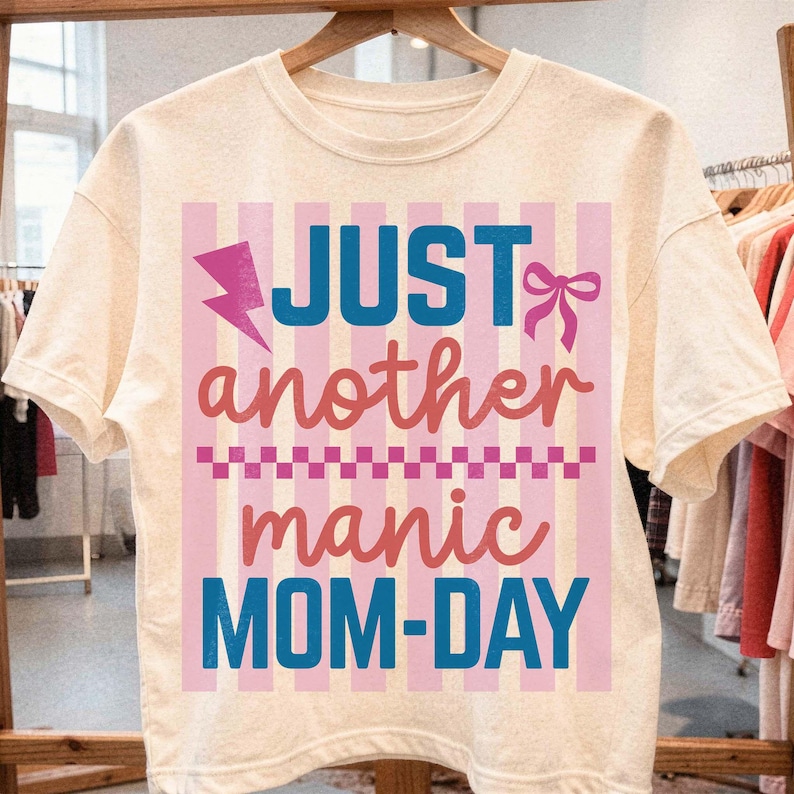 An ivory graphic t-shirt displayed on a wooden hanger in a bright, modern boutique. The shirt features a colorful motherhood design with the quote Just Another Manic Mom-Day in blue and coral typography over a pink striped backdrop. A pink lightning bolt and matching bow add a playful retro touch. This retail mockup emphasizes the high-quality, boutique feel of the t-shirt, making it an ideal visual for shoppers looking for a funny Mother’s Day gift or a trendy addition to their casual wardrobe.