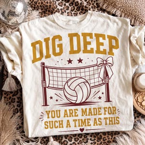 Op de afbeelding: Een crèmekleurig t-shirt met een bordeauxrode en gouden afbeelding. De tekst luidt DIG DEEP en YOU ARE MADE FOR SUCH A TIME AS THIS. Het ontwerp bevat een volleybalnet met een grote volleybal in het midden en een decoratieve strik aan de zijkant.