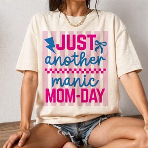 A lifestyle mockup of a woman sitting on a wooden ledge wearing an ivory oversized graphic tee with the quote Just Another Manic Mom-Day. The design features bold pink and blue script, a lightning bolt icon, and a decorative bow over a pink striped square. Styled with denim shorts and a gold chain necklace, this image highlights the shirt's relaxed, trendy fit. It is an excellent visual for showcasing the t-shirt as a stylish and relatable streetwear option for busy mothers.