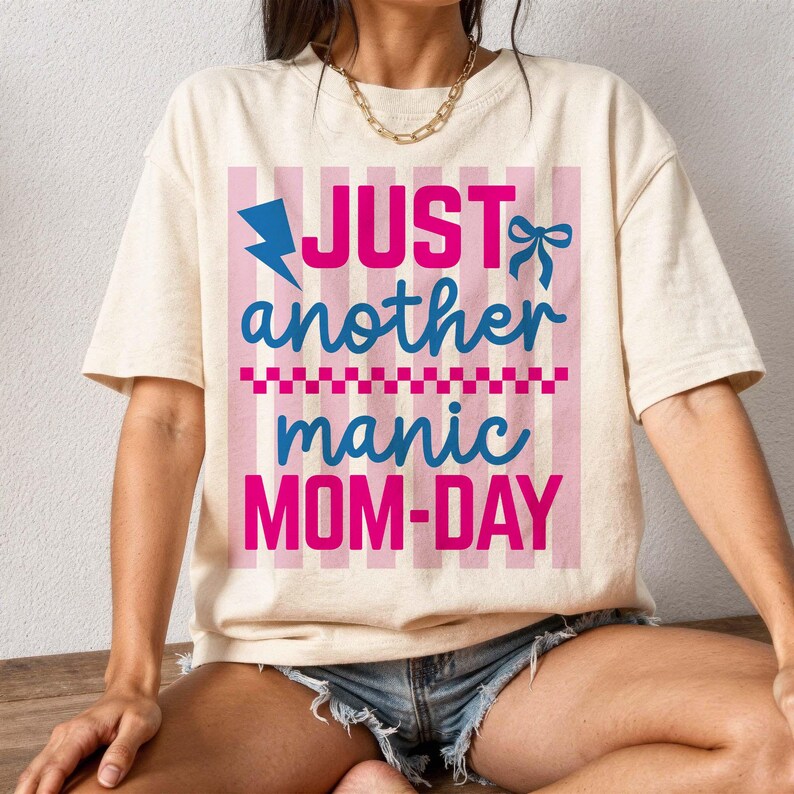 A lifestyle mockup of a woman sitting on a wooden ledge wearing an ivory oversized graphic tee with the quote Just Another Manic Mom-Day. The design features bold pink and blue script, a lightning bolt icon, and a decorative bow over a pink striped square. Styled with denim shorts and a gold chain necklace, this image highlights the shirt's relaxed, trendy fit. It is an excellent visual for showcasing the t-shirt as a stylish and relatable streetwear option for busy mothers.