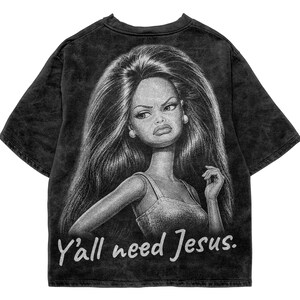 May include: A black acid-wash t-shirt featuring a large, detailed graphic of a doll with long dark hair and a sceptical facial expression. Below the graphic, the text "Y'all need Jesus." is printed in a white, stylised, handwritten-style font.