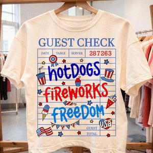以下が含まれることがあります： クリーム色のTシャツ。デザインは、ゲストチェックに似たグラフィックです。「Guest Check」、「Hot Dogs」、「Fireworks」、「Freedom」の文字と、星や旗などの愛国的なイラストが描かれています。