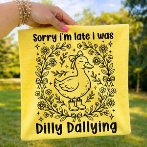 Puede incluir: Cojín amarillo con el texto "Sorry I'm late I was Dilly Dallying". Presenta un dibujo lineal negro de un pato con botas, rodeado de flores y lazos.