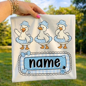 Puede incluir: Camiseta blanca con tres patos de dibujos animados que llevan sombreros y flotadores azules de lunares. Los patos están de pie sobre sus patas. Debajo, un diseño de cuadros vichy azul y blanco con la palabra "name" en negro.