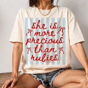 Könnte beinhalten: Cremefarbenes T-Shirt mit dem roten Schriftzug "she is more precious than rubies". Der Text befindet sich vor einem hellblau gestreiften Hintergrund mit weißen Punkten. Das Shirt hat kurze Ärmel.