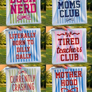 May include: Collection of light blue t-shirts with various phrases in bold, colorful fonts. Phrases include "BOOK NERD", "TIRED MOMS CLUB", "LITERALLY BORN TO DILLY DALLY", "TIRED TEACHERS CLUB", "CURRENTLY CRASHING OUT", and "MOTHER HOOD". Each shirt has a bow graphic.