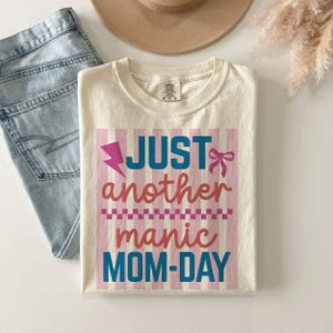A professional flat lay mockup featuring a folded ivory Comfort Colors t-shirt on a white surface. The shirt displays the Just Another Manic Mom-Day graphic with pink and blue accents over a striped background. Styled alongside light-wash denim jeans, a tan wide-brim felt hat, and a sprig of dried pampas grass, this aesthetic presentation emphasizes the t-shirt's versatility. It effectively shows how the shirt can be styled for a trendy, boho-chic mom outfit for the spring and summer seasons.