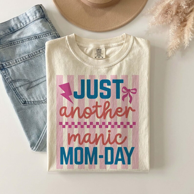 A professional flat lay mockup featuring a folded ivory Comfort Colors t-shirt on a white surface. The shirt displays the Just Another Manic Mom-Day graphic with pink and blue accents over a striped background. Styled alongside light-wash denim jeans, a tan wide-brim felt hat, and a sprig of dried pampas grass, this aesthetic presentation emphasizes the t-shirt's versatility. It effectively shows how the shirt can be styled for a trendy, boho-chic mom outfit for the spring and summer seasons.