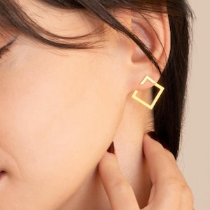 May include: A close-up of a person wearing a gold square earring. The earring is a simple, open square design. The person has dark hair and is touching their ear. The background is a solid, neutral color.