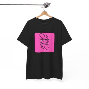 May include: Black t-shirt with a pink square graphic. The graphic features a stylized black design with the letters "EKB". The t-shirt is hanging on a wooden hanger.
