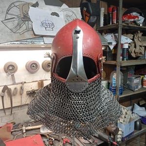 May include: A red medieval helmet with a silver face guard and a chainmail neck guard. The helmet is made of metal and leather. The background shows tools and equipment.