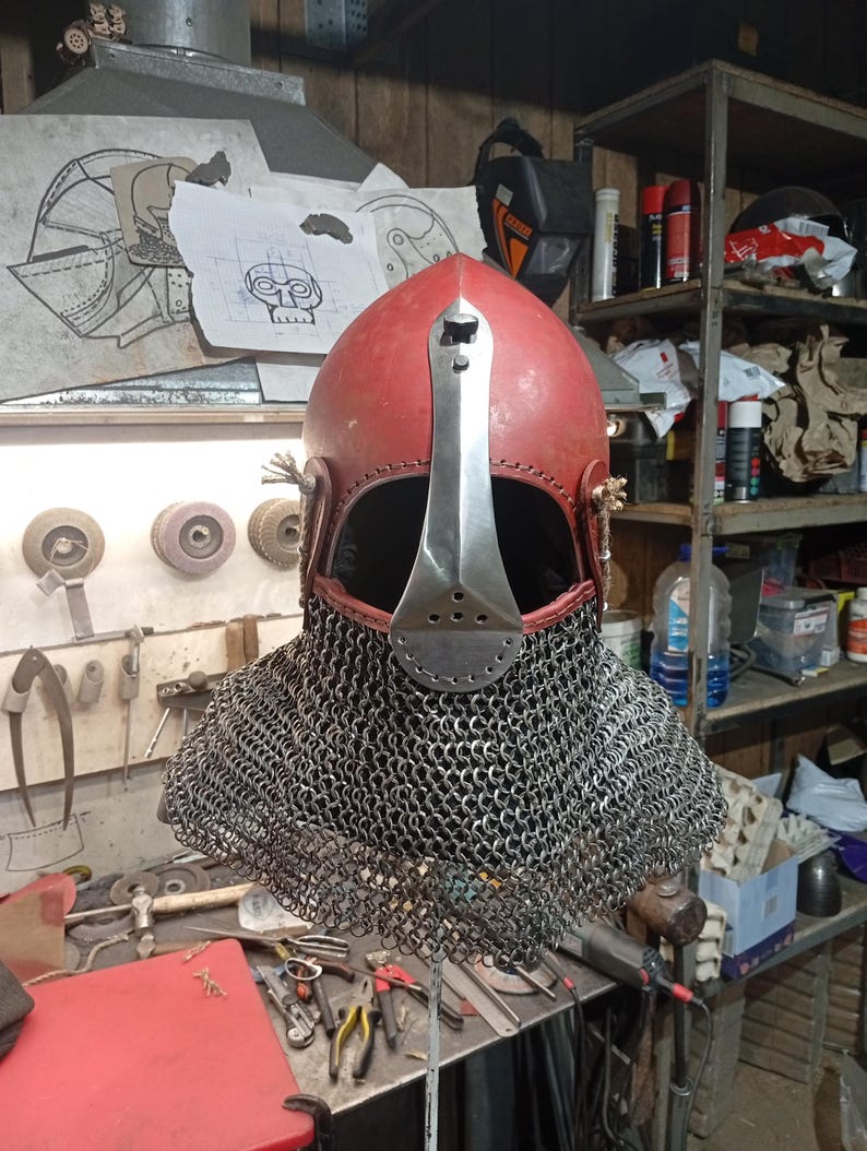May include: A red medieval helmet with a silver face guard and a chainmail neck guard. The helmet is made of metal and leather. The background shows tools and equipment.