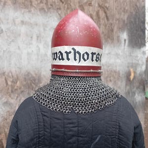 May include: A red and white medieval helmet with the word "warhorse" printed on the front. The helmet is worn over a chainmail collar and a black quilted garment.