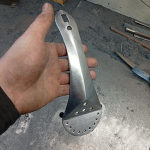 May include: A hand-forged, polished metal tool with a long, tapered handle and a flared, perforated head. The tool has a square hole and a circular hole near the top of the handle. The metal has a brushed finish.