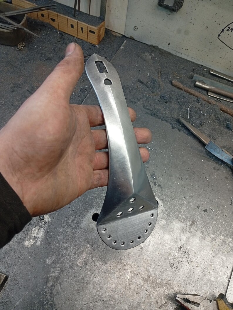 May include: A hand-forged, polished metal tool with a long, tapered handle and a flared, perforated head. The tool has a square hole and a circular hole near the top of the handle. The metal has a brushed finish.
