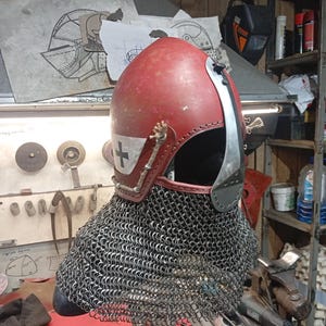 May include: A medieval-style helmet with a red, leather-covered top and a silver face guard. The helmet features a white cross emblem and is attached to a chainmail coif. The design suggests historical reenactment or costume use.