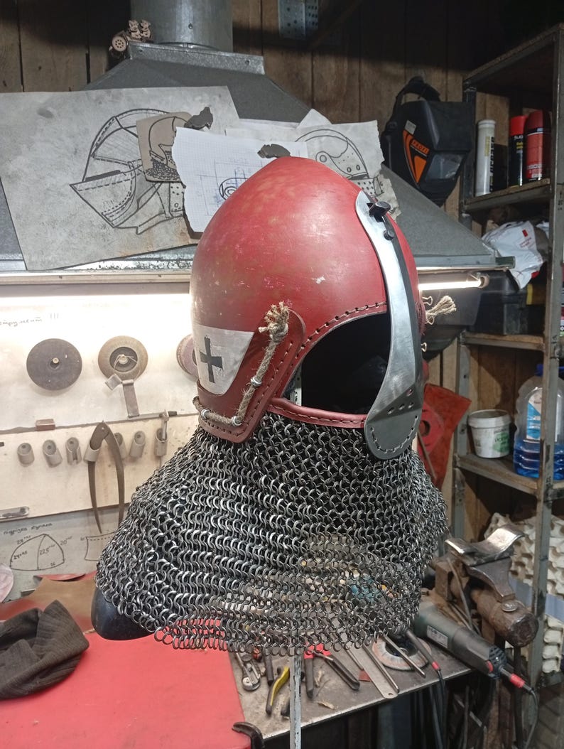May include: A medieval-style helmet with a red, leather-covered top and a silver face guard. The helmet features a white cross emblem and is attached to a chainmail coif. The design suggests historical reenactment or costume use.