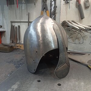 May include: A silver-colored metal helmet with a curved visor, likely for historical reenactment or display. The helmet has a rounded top and is made of a single piece of metal. It sits on a metal surface in a workshop setting.