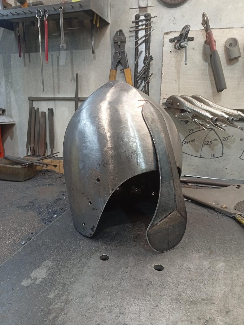 May include: A silver-colored metal helmet with a curved visor, likely for historical reenactment or display. The helmet has a rounded top and is made of a single piece of metal. It sits on a metal surface in a workshop setting.