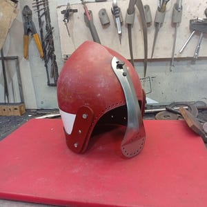 May include: A red medieval helmet with a silver visor and white accents. The helmet is made of metal and leather, with rivets securing the pieces. It sits on a red surface, with tools visible in the background.