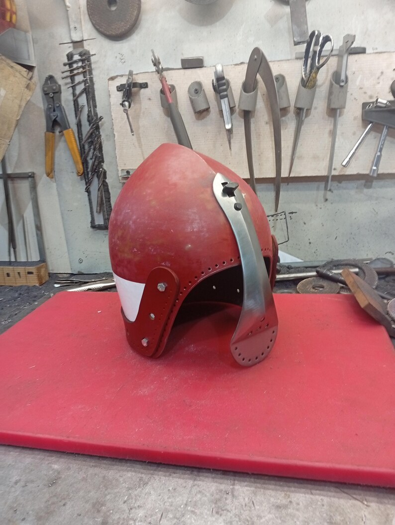 May include: A red medieval helmet with a silver visor and white accents. The helmet is made of metal and leather, with rivets securing the pieces. It sits on a red surface, with tools visible in the background.
