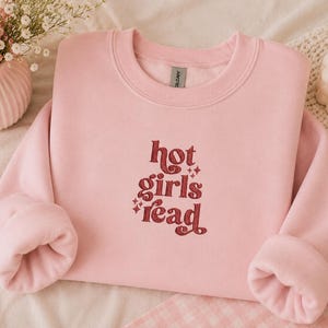 Op de afbeelding: Een lichtroze sweatshirt met de tekst "hot girls read" in bordeauxrood, in een retro-stijl lettertype. De sweatshirt heeft een ronde hals en lange mouwen. De afbeelding bevat ook een roze vaas met bloemen en een brandende kaars.