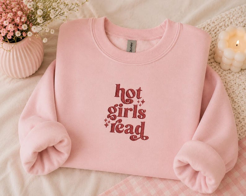Op de afbeelding: Een lichtroze sweatshirt met de tekst "hot girls read" in bordeauxrood, in een retro-stijl lettertype. De sweatshirt heeft een ronde hals en lange mouwen. De afbeelding bevat ook een roze vaas met bloemen en een brandende kaars.