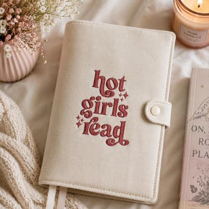 Op de afbeelding: Cr&egrave;mekleurige boekomslag met de geborduurde tekst "hot girls read" in een roodachtig bruine kleur. De omslag heeft een knoopsluiting en twee witte lint-bladwijzers. Een brandende kaars en een vaas met bloemen staan op de achtergrond.