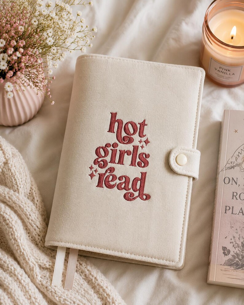 Op de afbeelding: Cr&egrave;mekleurige boekomslag met de geborduurde tekst "hot girls read" in een roodachtig bruine kleur. De omslag heeft een knoopsluiting en twee witte lint-bladwijzers. Een brandende kaars en een vaas met bloemen staan op de achtergrond.