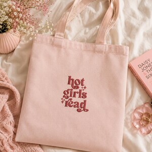 Op de afbeelding: Een lichtroze draagtas met de geborduurde tekst "hot girls read" in bordeauxrood. De tas heeft bijpassende roze handvatten. Een boek en bloemen staan op de achtergrond, geplaatst op een wit oppervlak.