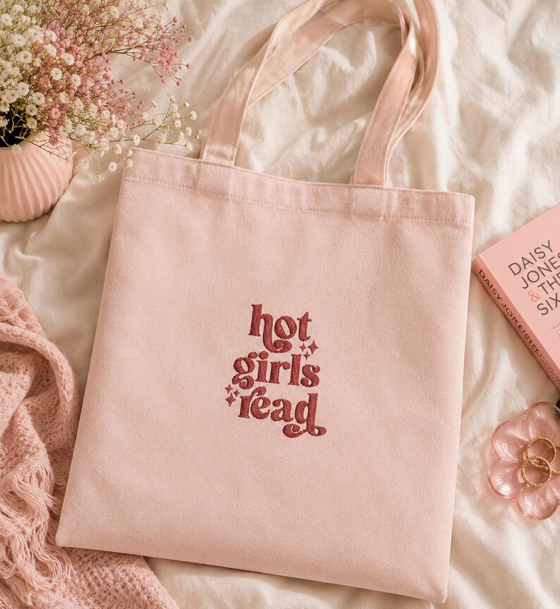 Op de afbeelding: Een lichtroze draagtas met de geborduurde tekst "hot girls read" in bordeauxrood. De tas heeft bijpassende roze handvatten. Een boek en bloemen staan op de achtergrond, geplaatst op een wit oppervlak.