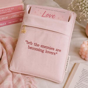 May include: A light pink book sleeve with a front pocket and a gold zipper. The text "*brb the enemies are becoming lovers*" is embroidered on the front. A book with the title "Love" is inside the sleeve.
