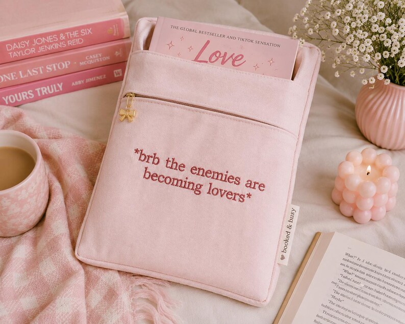 May include: A light pink book sleeve with a front pocket and a gold zipper. The text "*brb the enemies are becoming lovers*" is embroidered on the front. A book with the title "Love" is inside the sleeve.
