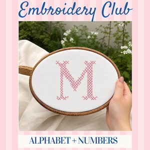 May include: An oval wooden embroidery hoop containing white fabric with a pink cross-stitched letter M. The text Embroidery Club and ALPHABET + NUMBERS appear in blue at the top and bottom of the image against a light pink striped background.