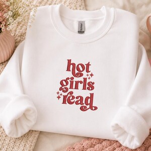 Op de afbeelding: Witte crewneck sweatshirt met de tekst "hot girls read" in rode letters. De sweatshirt heeft lange mouwen en een relaxte pasvorm. De tekst is in een retro lettertype met steraccenten.