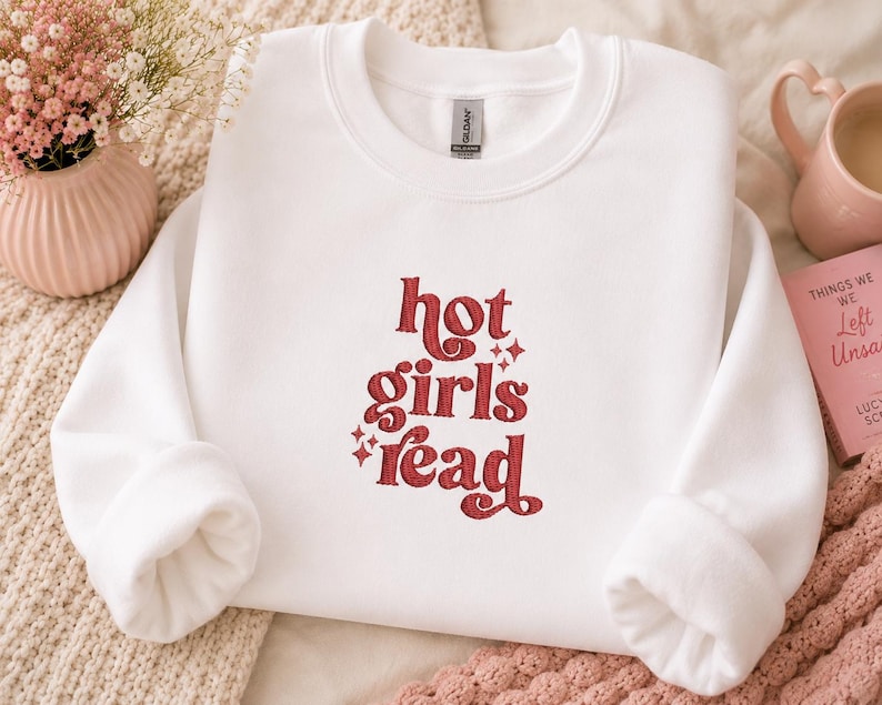 Op de afbeelding: Witte crewneck sweatshirt met de tekst "hot girls read" in rode letters. De sweatshirt heeft lange mouwen en een relaxte pasvorm. De tekst is in een retro lettertype met steraccenten.