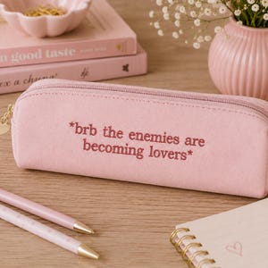 May include: A light pink pencil case with the text "*brb the enemies are becoming lovers*" embroidered on it. The case has a gold zipper and heart-shaped charm. Two pens and a notepad are also visible.