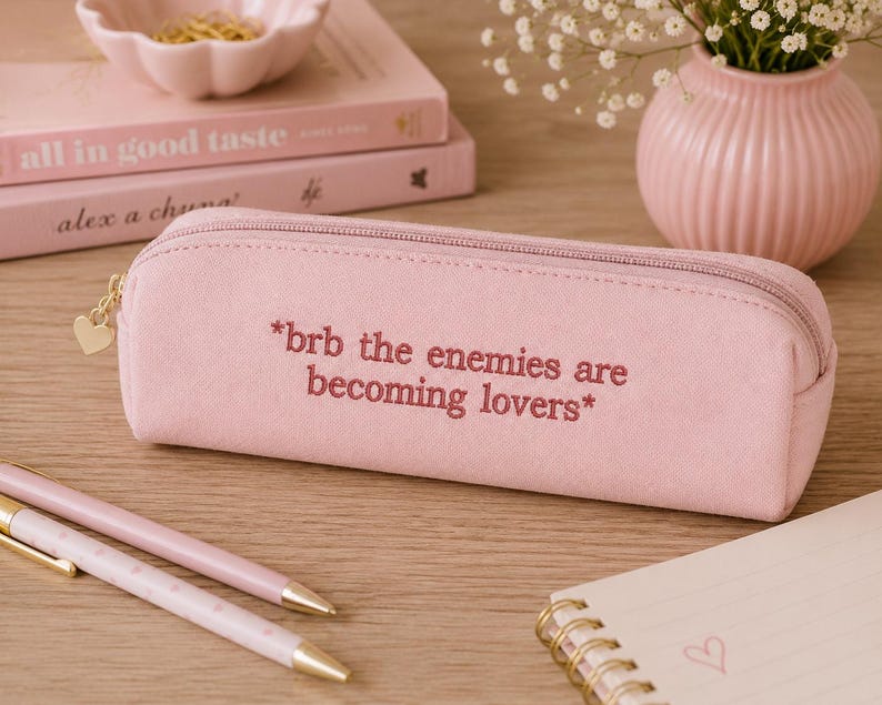 May include: A light pink pencil case with the text "*brb the enemies are becoming lovers*" embroidered on it. The case has a gold zipper and heart-shaped charm. Two pens and a notepad are also visible.