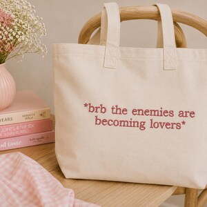 May include: Cream-colored tote bag with the embroidered text "*brb the enemies are becoming lovers*". The bag has two handles and is sitting on a wooden chair. A pink vase with flowers and books are in the background.