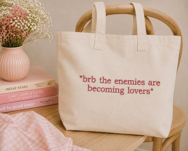 May include: Cream-colored tote bag with the embroidered text "*brb the enemies are becoming lovers*". The bag has two handles and is sitting on a wooden chair. A pink vase with flowers and books are in the background.