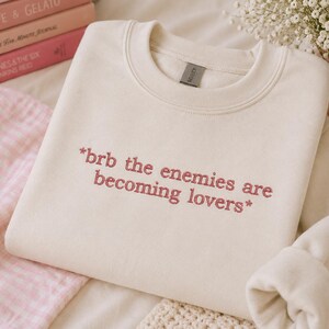 May include: Cream-colored sweatshirt with the embroidered text "*brb the enemies are becoming lovers*". The crewneck sweatshirt is folded, with the sleeves visible. A stack of pink books and a small vase with white flowers are in the background.